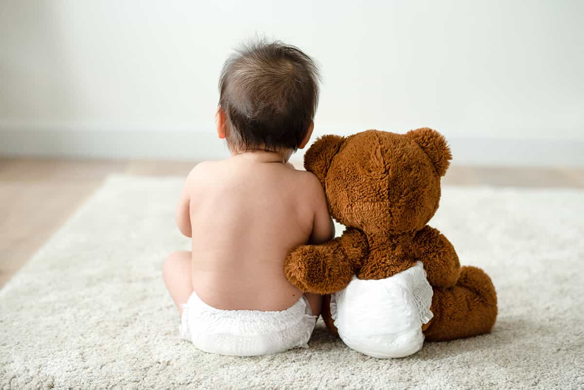 Baby sitting facing away from the camera next to a teddy bear that has its arm around the baby's back